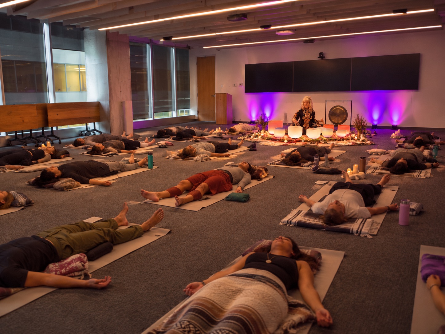 Chris Young-Ginzburg leading a group sound bath with crystal bowls and a gong for a team resting on mats in an office